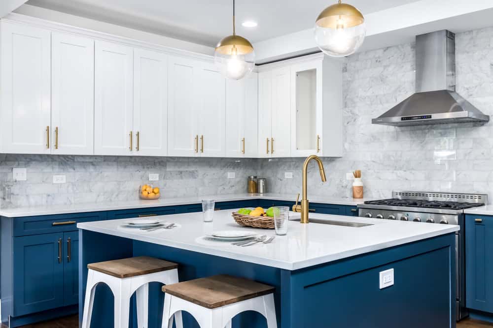 Modern kitchen with a white marble backsplash, blue lower cabinets, and white upper cabinets. A stainless steel hood and appliances are present, including those eligible for Refrigerator Repair Cook County services. The island has a white countertop, sink, brass faucet, three stools, and pendant lights above.