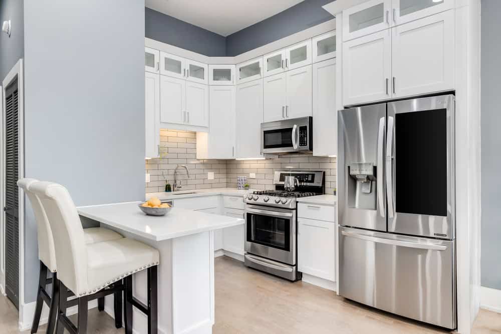 Modern kitchen with white cabinets, stainless steel appliances, and a light wood floor. Features a French door refrigerator and built-in microwave. The backsplash has white subway tiles. An island with two white cushioned chairs is in the foreground—ideal for those who appreciate quality workmanship like Dishwasher Repair Cook County.